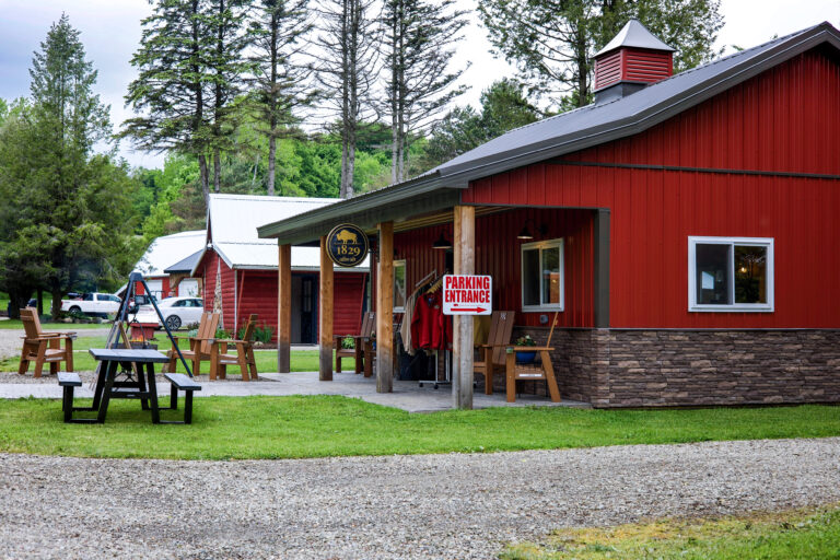 A rustic red building with a stone foundation, picnic tables on a grassy lawn, trees in the background, and a "Parking Entrance" sign hanging near the covered porch.