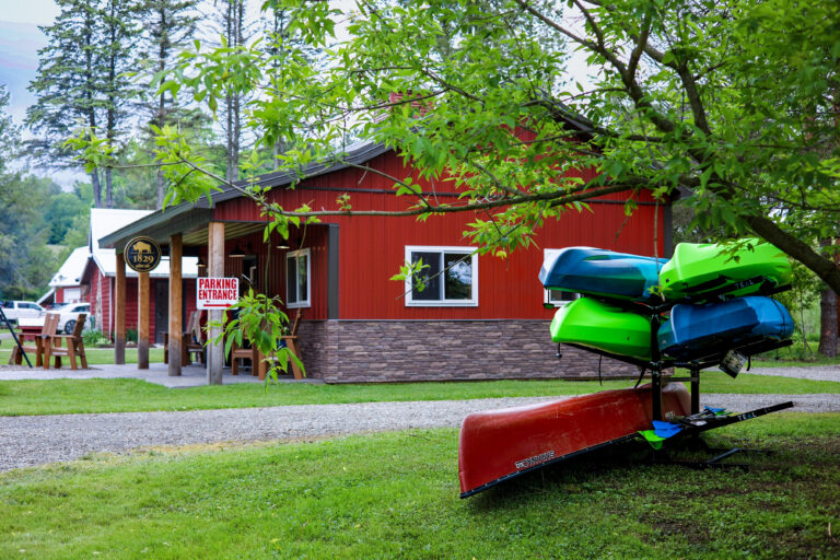 A red building with a "Parking Entrance" sign sits behind a tree. In the foreground, colorful kayaks and canoes are stacked on a grassy area.