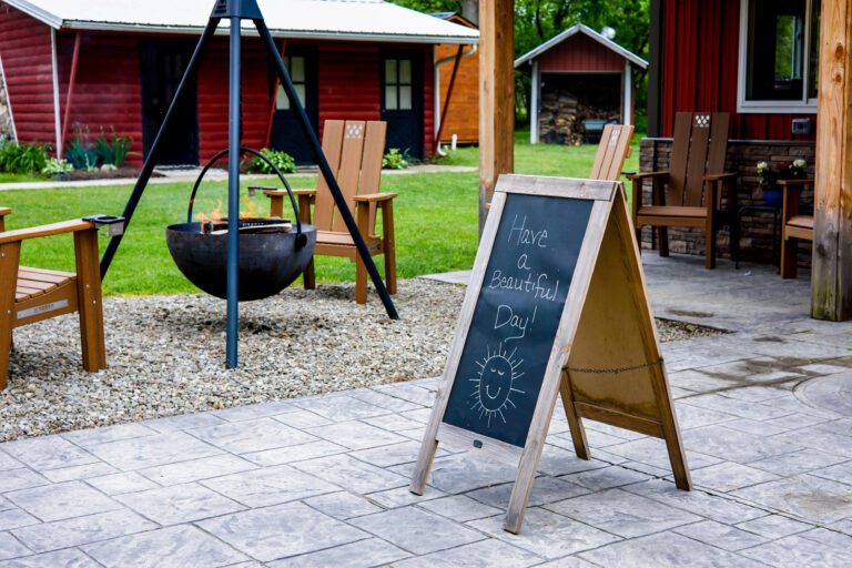 A chalkboard sign reads "Have a Beautiful Day!" with a drawing of a sun. It stands on a patio with wooden chairs, a fire pit, and a red cabin in the background.