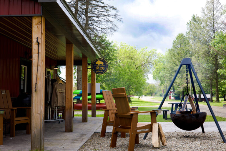 A covered porch with wooden chairs and clothing for sale stands beside a campfire pit on a tripod, with trees, kayaks, and a campground road visible in the background under a partly cloudy sky.